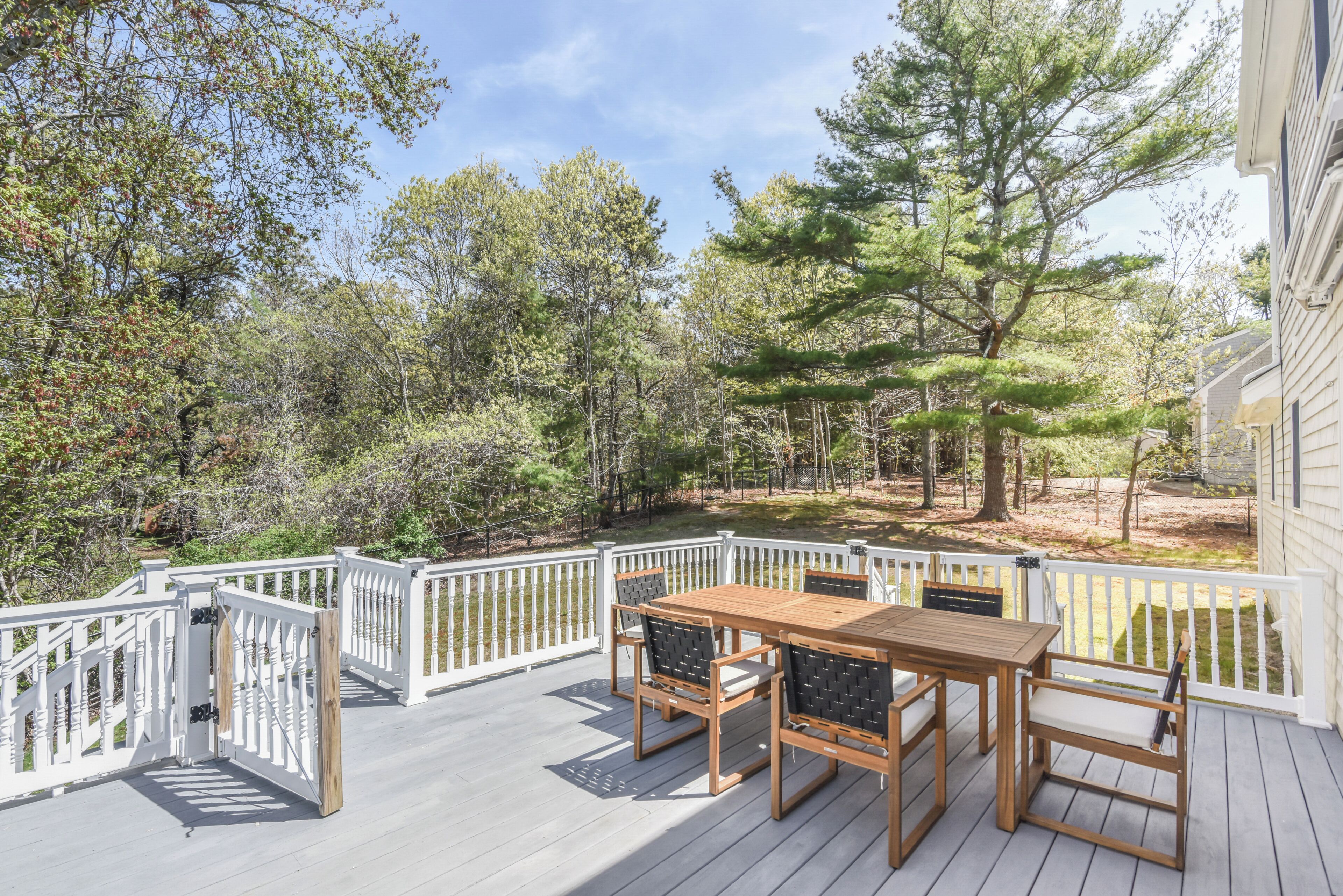 A wooden deck with a table and chairs, surrounded by white railings and trees.