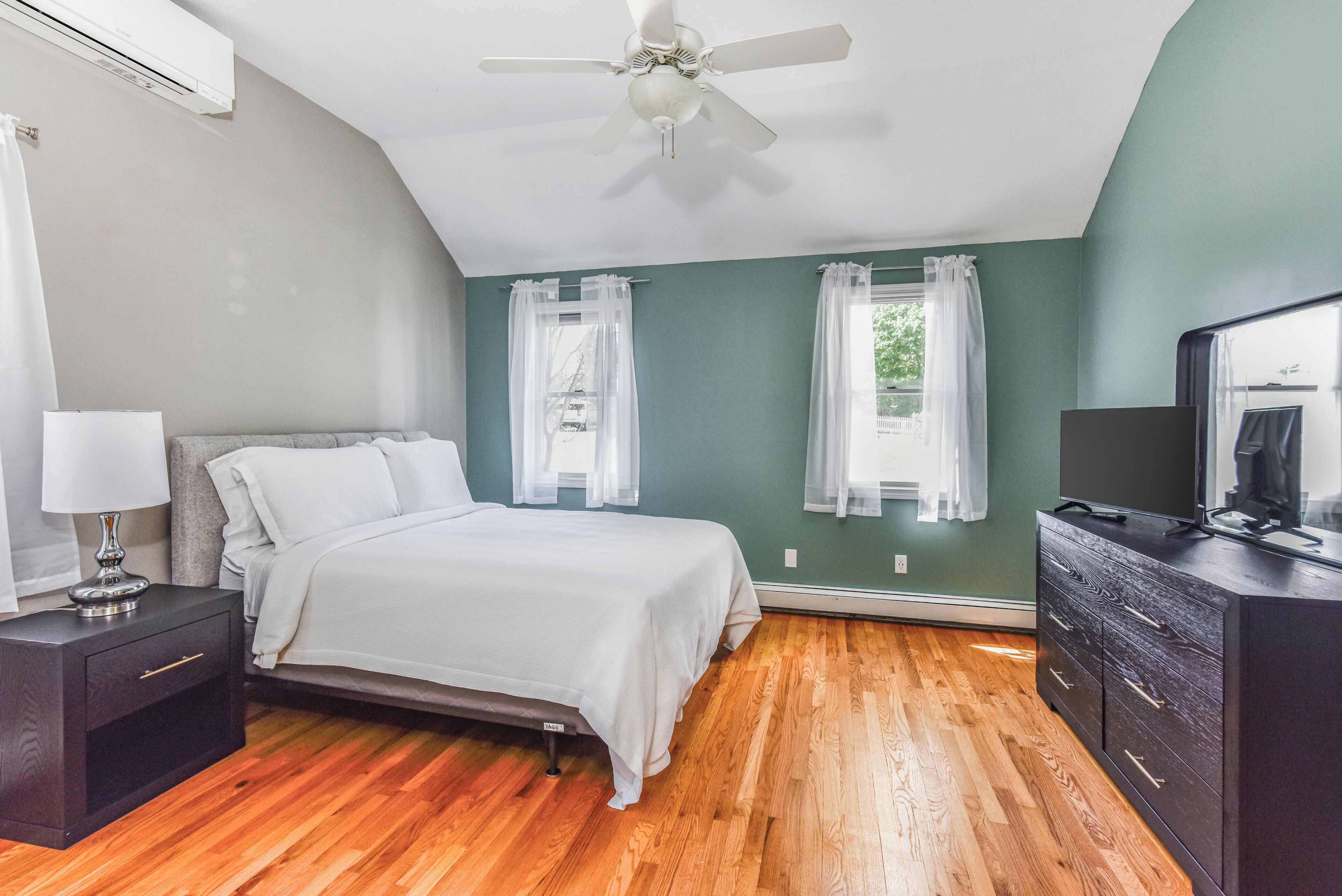 A bedroom with a bed, a nightstand, a dresser, a ceiling fan, and two windows with curtains.