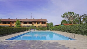 Una piscina rectangular con plataforma de madera, rodeada de setos y árboles, con un edificio al fondo.
