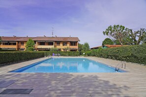 Ein rechteckiger Swimmingpool mit einer Holzterrasse, umgeben von Hecken und Bäumen, mit einem Gebäude im Hintergrund.
