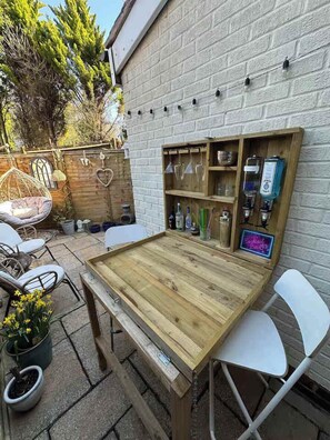 Ein Holztisch im Freien mit Stühlen, einer hängenden Lampe und einem Regal mit Flaschen und Gläsern.