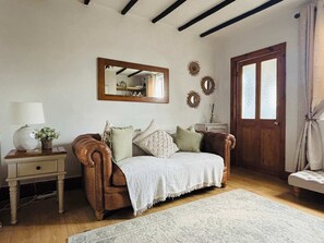 Ein gemütliches Zimmer mit braunem Ledersofa, einer weißen Tagesdecke, Holzboden, Holztür und einem Spiegel an der Wand.