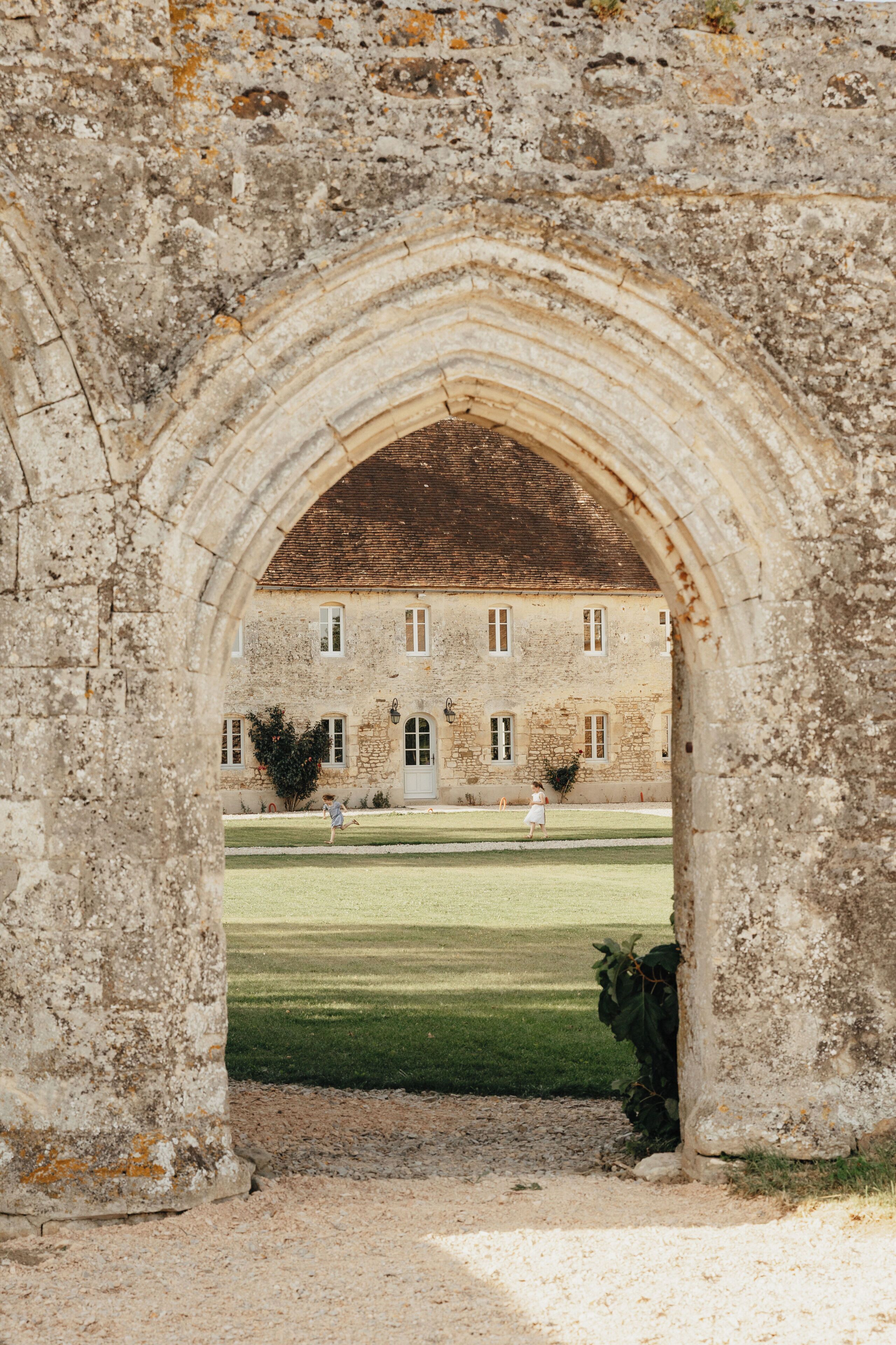 An arched stone gateway leading to a historic building with a symmetrical facade, multiple windows, and a well-maintained lawn.