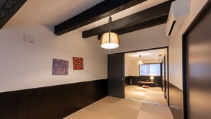 Un pasillo con suelo de tatami, una lámpara colgante y una puerta que da a una habitación con ventana y mesita de noche.