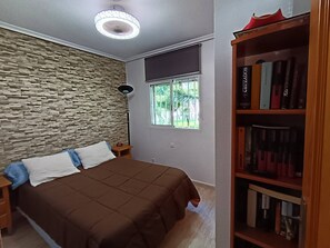 Une chambre avec un mur en pierre, un lit recouvert d’une couverture marron, une table de chevet, une lampe sur pied, une fenêtre donnant sur la verdure et une bibliothèque remplie de livres.