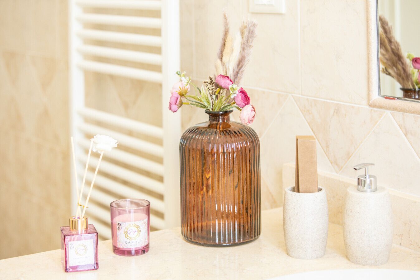A bathroom vanity with a vase of flowers, a pink candle, a soap dispenser, and a small bottle.