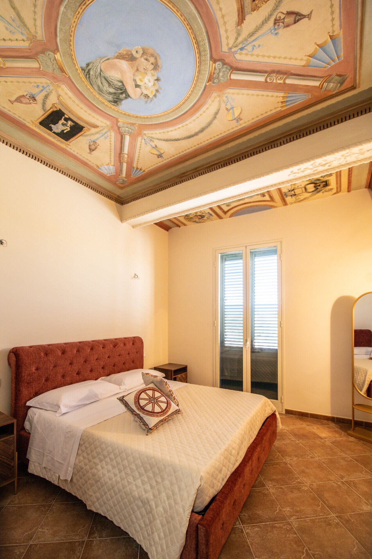 A bedroom with a large bed, a red upholstered headboard, a ceiling with frescoes, and a sliding glass door.