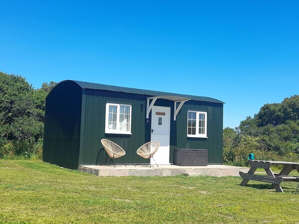 A small, dark green building with two windows, a door, and a bench outside.