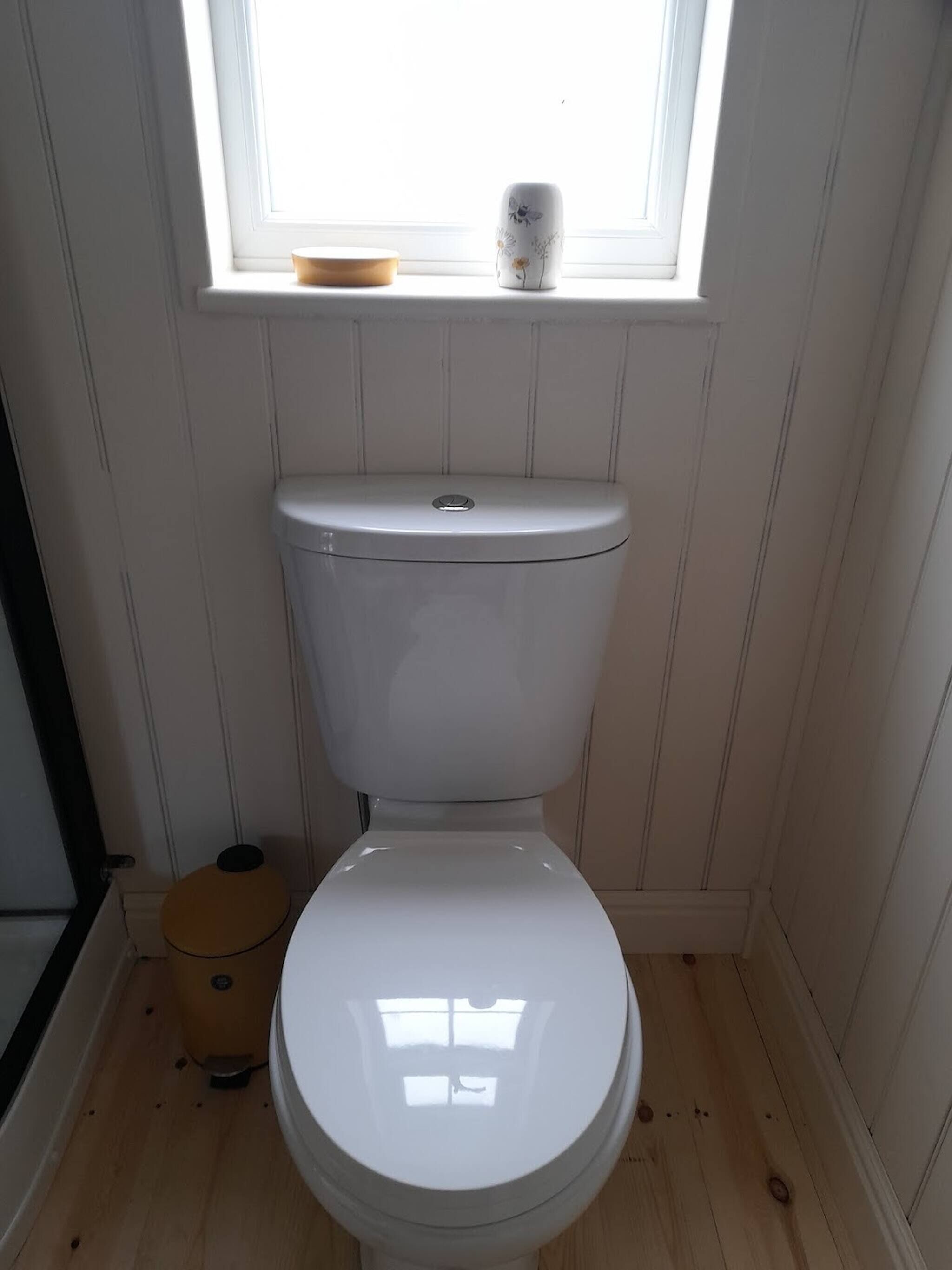 Een wit toilet met een raam erboven, een klein houten krukje en een prullenbak.