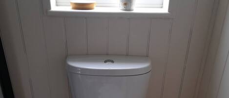 Een wit toilet met een raam erboven, een klein houten krukje en een prullenbak.