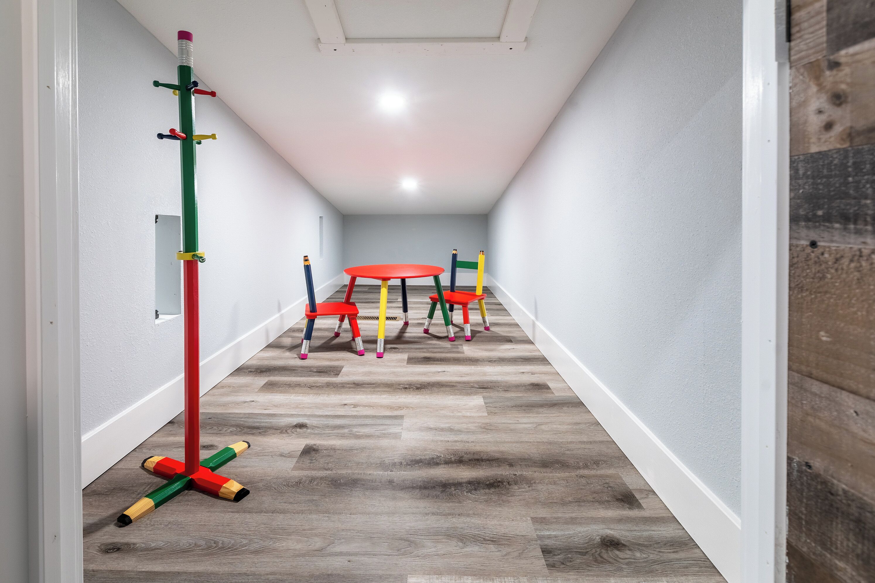 Un pasillo estrecho con piso de madera, una mesa roja y sillas de colores.