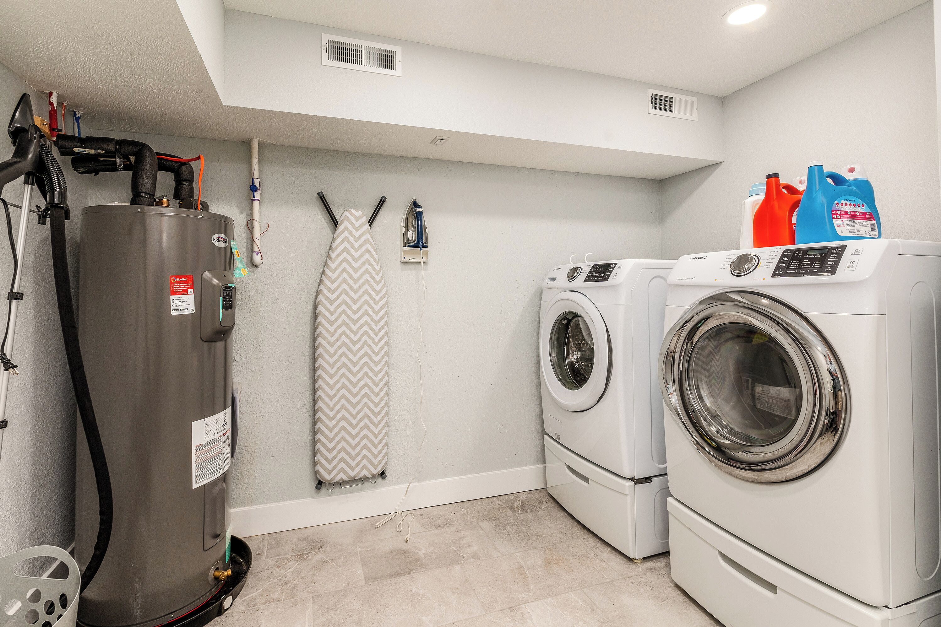 A laundry room with a water heater, two washing machines, and a vent.