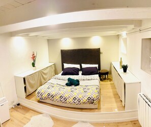 A modern bedroom with a bed, bedside tables, a small stool, and a potted plant.