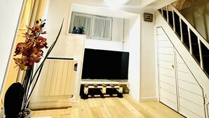 Huoneessa on puulattia, jalustalla oleva televisio, portaikko ja ikkuna, jossa on verhot.