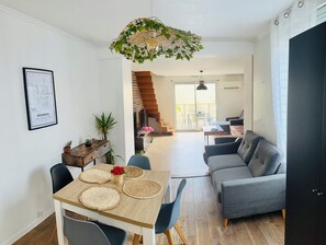 A modern living room with a grey sofa, a wooden dining table with woven placemats, a hanging light fixture, and a staircase leading to an upper level. - Cosy house near Paris (Colombes)