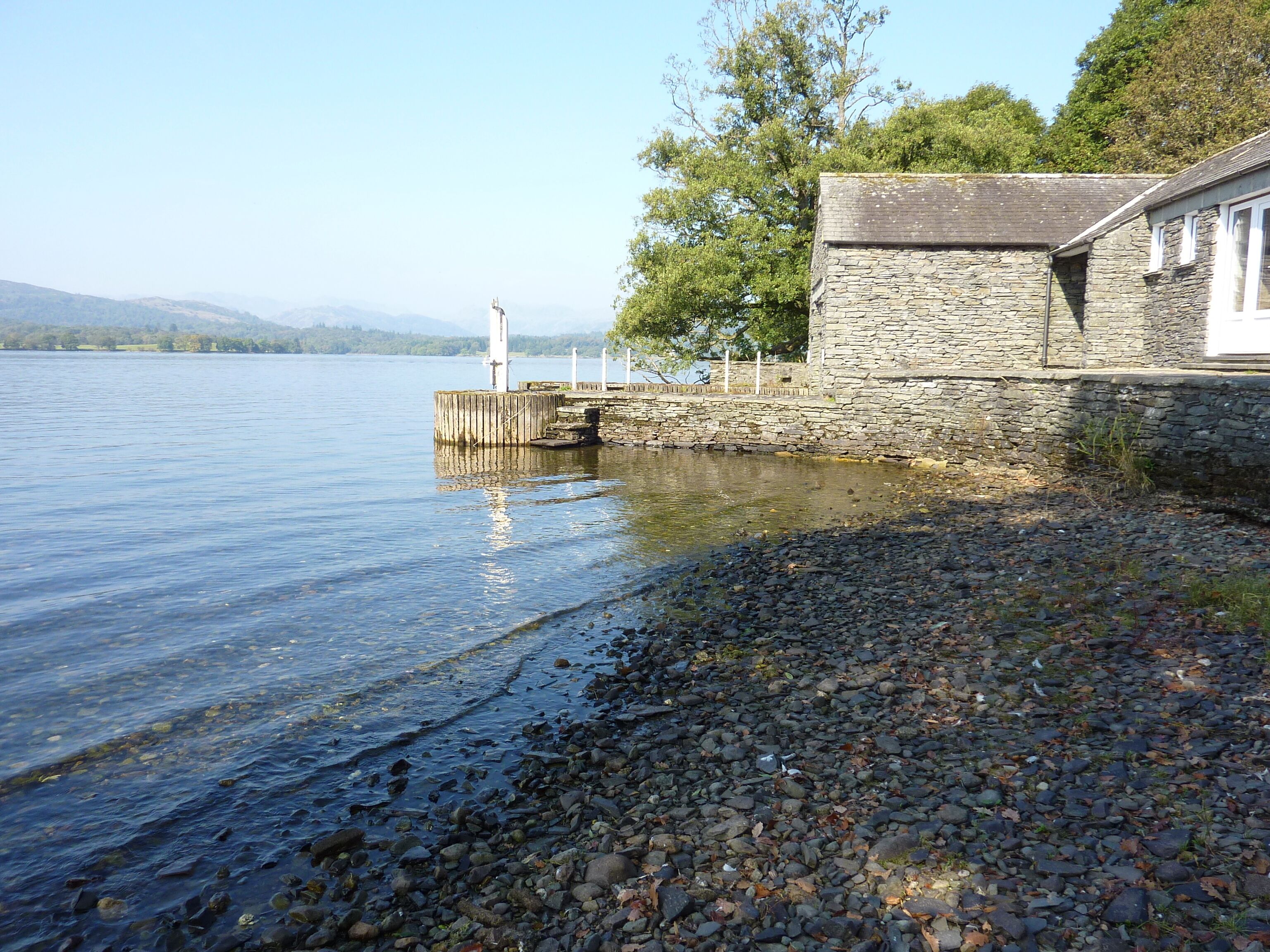 A stone building with a dock extending into calm water, surrounded by trees and distant hills.