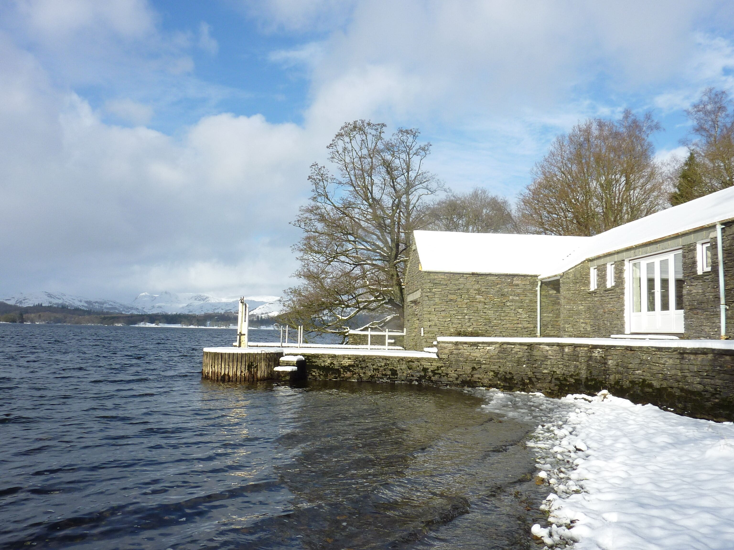 A stone building with a snow-covered roof, a dock extending into a lake, and snow on the ground.