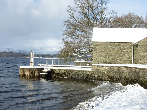 A stone pier extending into a lake with snow on the ground and a stone building in the background.