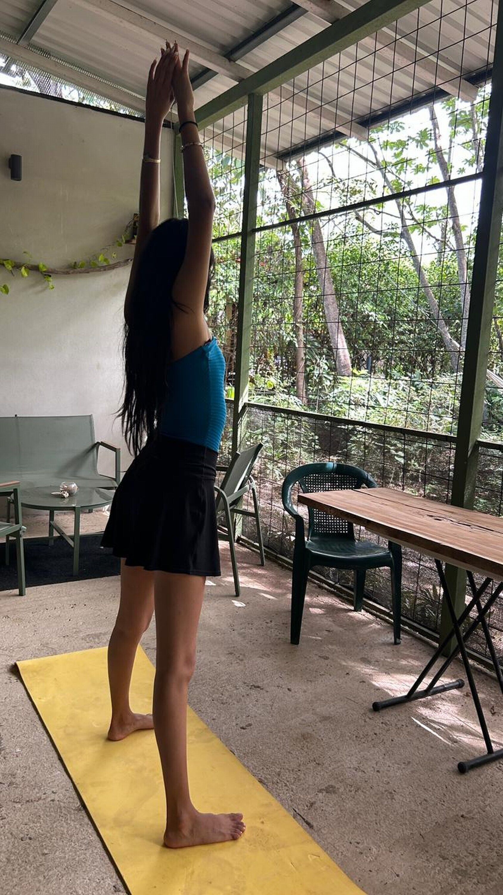 A person performing a yoga pose on a mat in a covered outdoor area with chairs and a table.