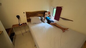 A person lying on a bed in a room with a wooden headboard, a bedside table with a lamp, and a window with curtains. - Cozy forest home one block from renowned Playa Grande surfing beach (Playa Grande)