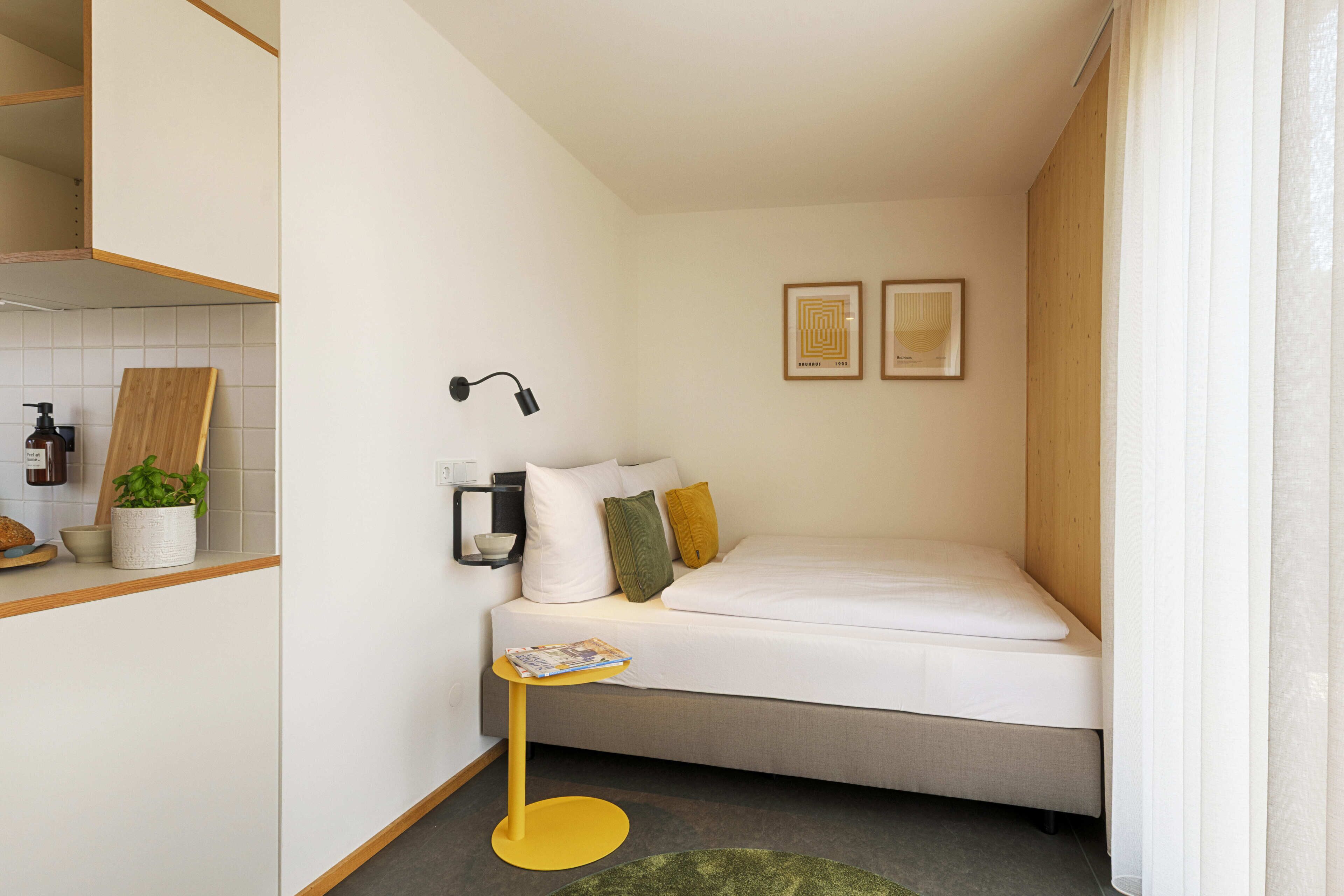 A compact hotel room with a bed, a small table, a wall-mounted lamp, and a potted plant.