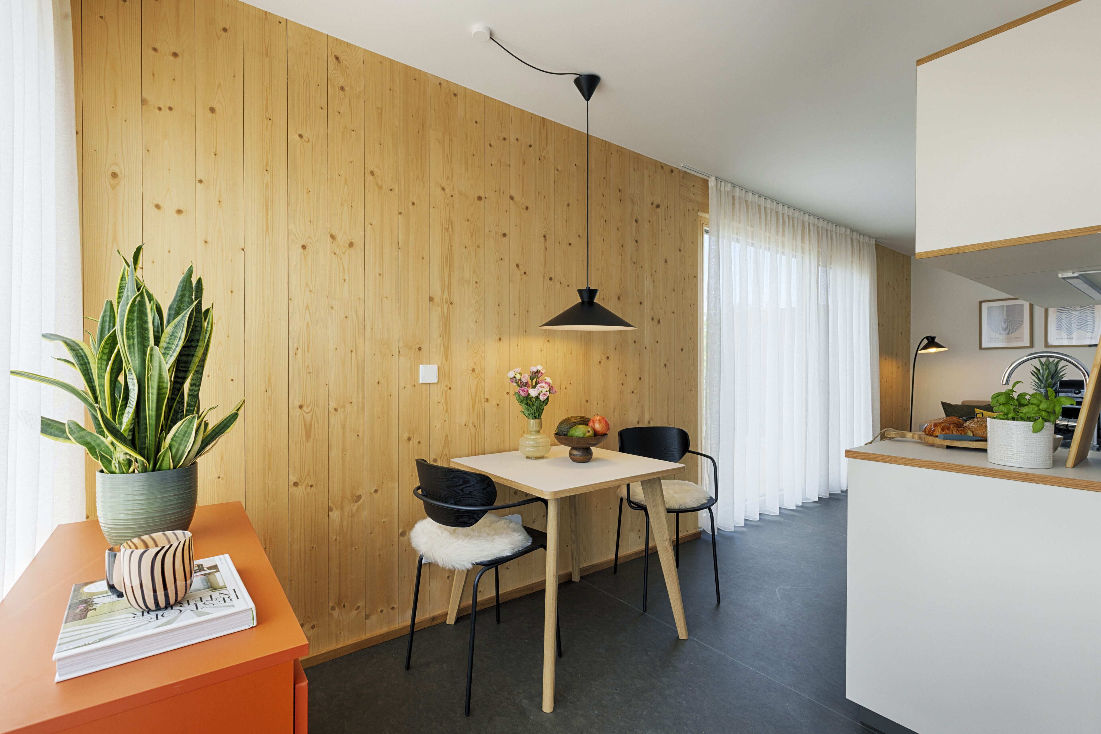 Een moderne keuken met houten lambrisering, een eethoek met een witte tafel, zwarte stoelen en een hangende hanglamp.