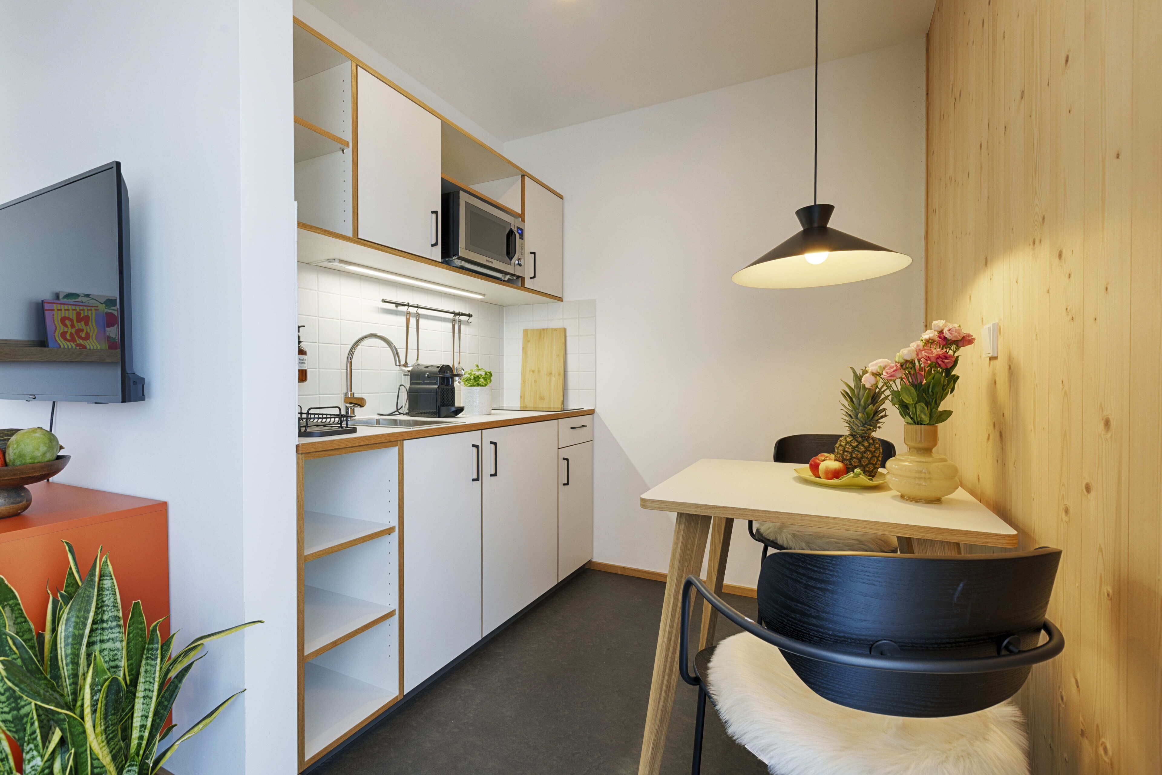 A compact kitchen with a microwave, toaster, and wooden cabinets. There's a dining table with a chair and a hanging light fixture.