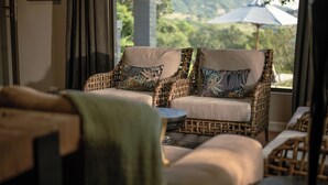 A wicker sofa with patterned cushions, a small round table, and a view of greenery through large windows.