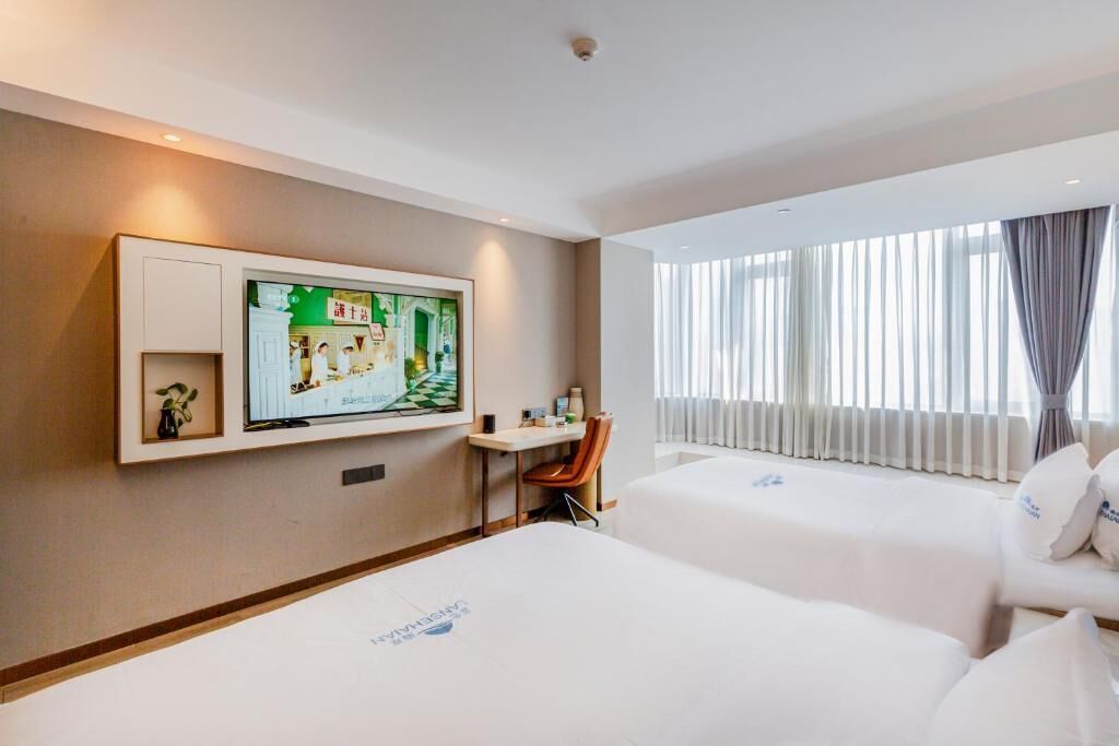 A hotel room with two beds, a desk, a chair, a TV mounted on the wall, and a window with curtains.
