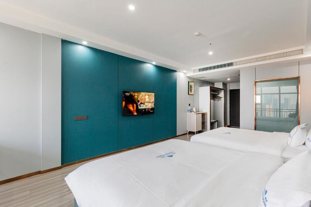 A hotel room with a teal accent wall, a flat-screen TV, two beds, a desk, and a window with a city view.