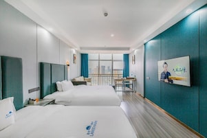 A hotel room with two beds, a desk, a chair, a window with a city view, and a mounted picture on the wall.