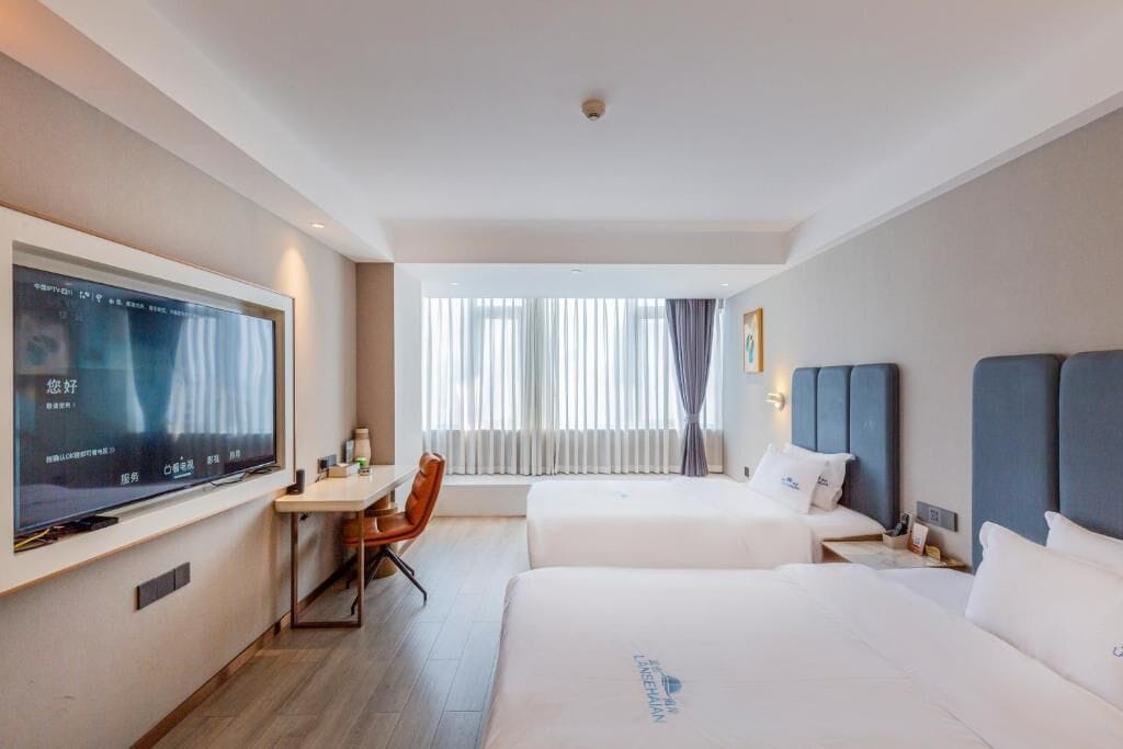 A hotel room with two beds, a desk, a chair, a TV, and a window with curtains.