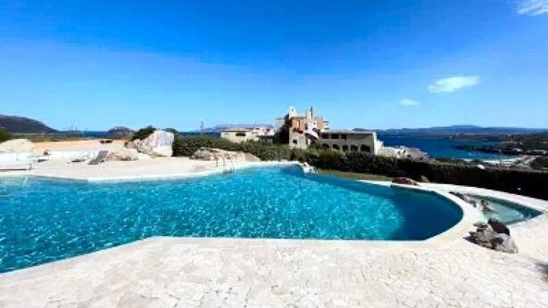 A swimming pool with a curved edge, a building with a white facade, and a clear blue sky. - PRESTIGE SHERDEN (Golfo Aranci)