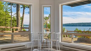 Une pièce dotée de grandes fenêtres donnant sur un lac, d’une terrasse en bois et d’une table blanche avec des chaises.