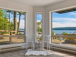 A room with large windows overlooking a lake, a wooden deck, and a white table with chairs.