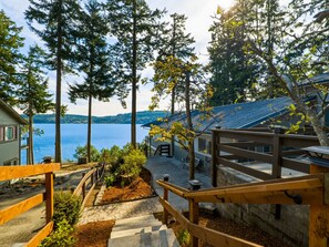 A wooden staircase leading to a deck overlooking a lake, surrounded by tall trees and a house with a blue roof. - Discovery Bay Beachview Escape in Port Townsend (Port Townsend)