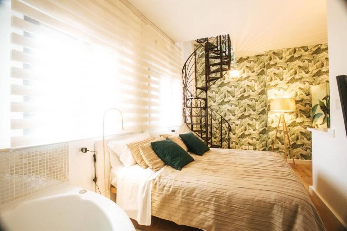A bedroom with a bed, a window with blinds, a wall-mounted light, and a spiral staircase.