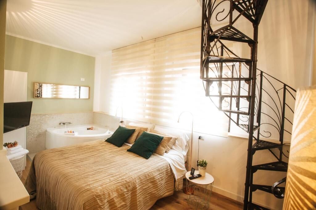 Un dormitorio con cama, mesita de noche, lámpara, televisor, ventana con persianas y una escalera.