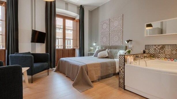 Una habitación de hotel moderna con cama, una zona de estar con sillón, un ventanal grande, televisor de pared y baño con bañera.