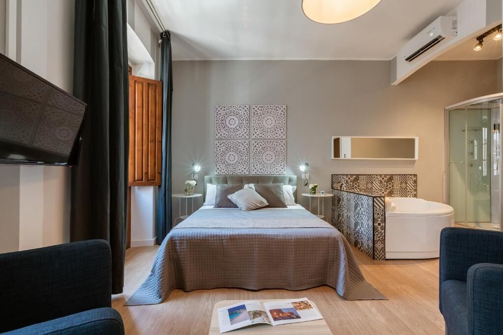 Ein modernes Hotelzimmer mit einem großen Bett, Nachttischen, einem integrierten Fernseher, einer Duschkabine aus Glas und einer kleinen Sitzecke.