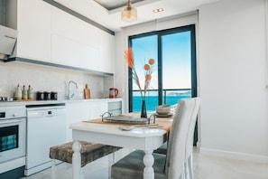A modern kitchen with white cabinetry, a dining area with a white table, and a view of the sea through a large window. - Gold Sand Suites (Alanya)
