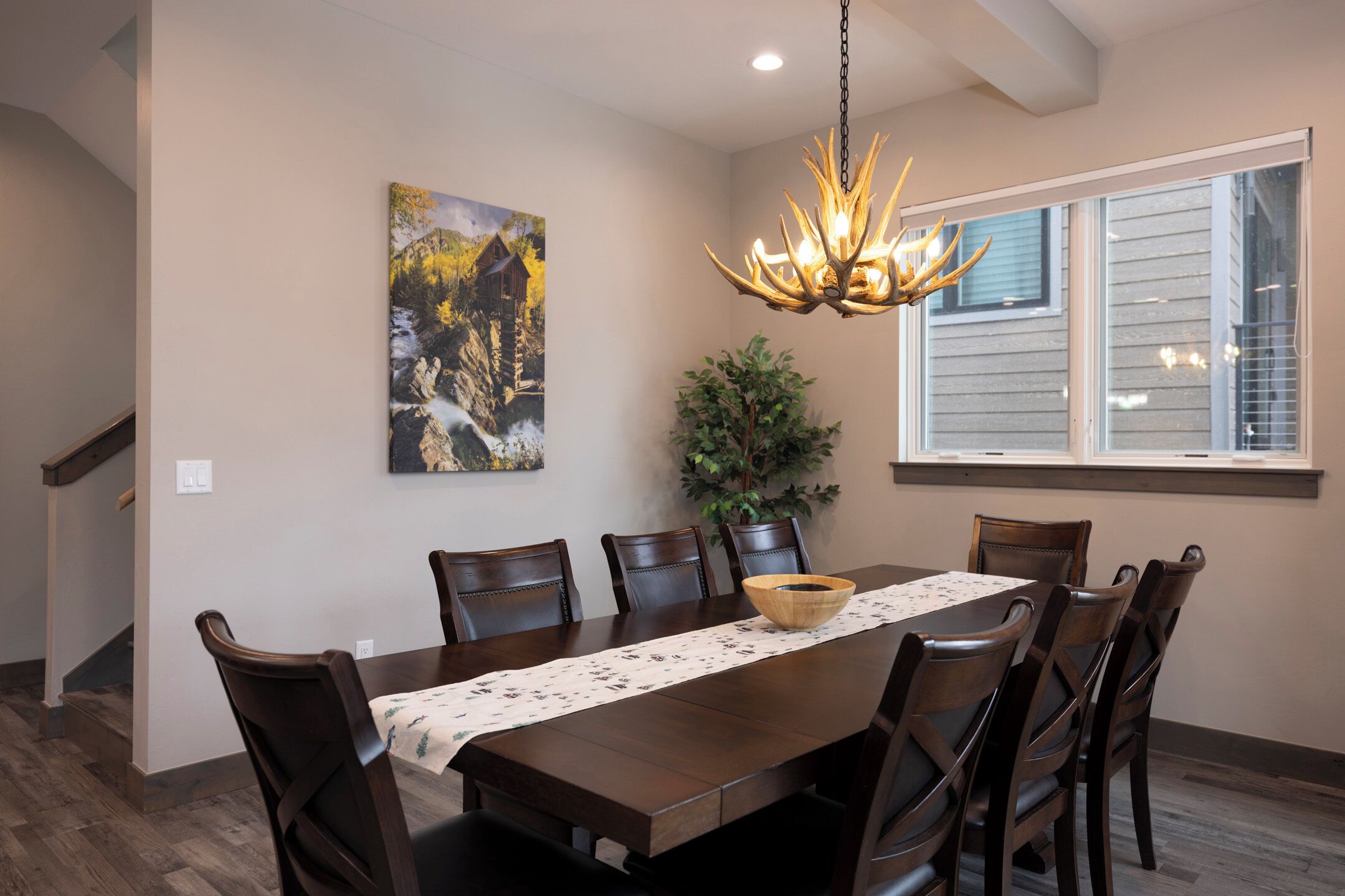 A dining room with a wooden table, chairs, a chandelier, a painting, and a window.