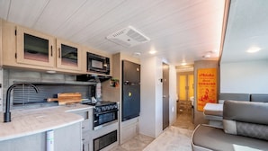 A compact kitchen with a microwave, stove, and refrigerator, a seating area with a sofa, and a door with a Hawaiian-themed sign. - Glamping Trailer Lanikai (Awaji)