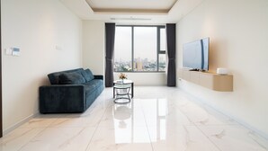 A modern living room with a sofa, a small round table with a vase, a flat-screen TV mounted on the wall, and a city view through large windows. - Imperial Suites by Kieu by Kita (Ho Chi Minh City)