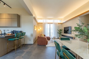 Eine moderne Küche mit Barhocker, einem Esstisch und einer Couch in einem hellen Wohnbereich mit großem Fenster und einer Pflanze.