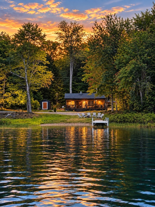 Una cabaña junto al lago con un muelle, rodeada de árboles y reflejando el atardecer.