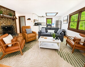 A cozy living room with a fireplace, a sofa, a coffee table, and a kitchen in the background. - Cozy Log Cabin by Phillips Lake, Pet Friendly, Near Acadia & Bangor (Dedham)