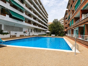 Una piscina con plataforma de baldosas, un área de descanso con banco y sombrilla, y un edificio de varios pisos con balcones y vegetación.