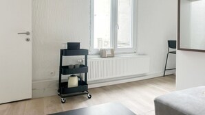 A room with a white door, a window with blinds, a radiator, a black cart with items, and a grey ottoman. - Charming Studio Steps from City Center (Ghent)