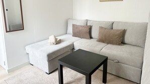 A neutral-colored sofa with two pillows, a small black table, a wall-mounted mirror, and a textured wall art piece. - Charming Studio Steps from City Center (Ghent)